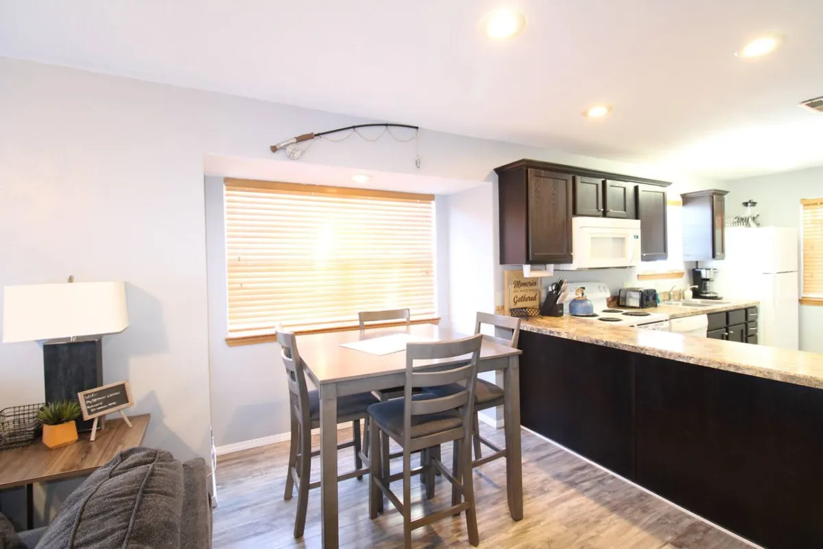 A cozy dining area featuring a dining table adjacent to a kitchen counter and a window with blinds. The table is ready for family meals or board games, and the open layout links the dining and cooking spaces, promoting shared experiences.