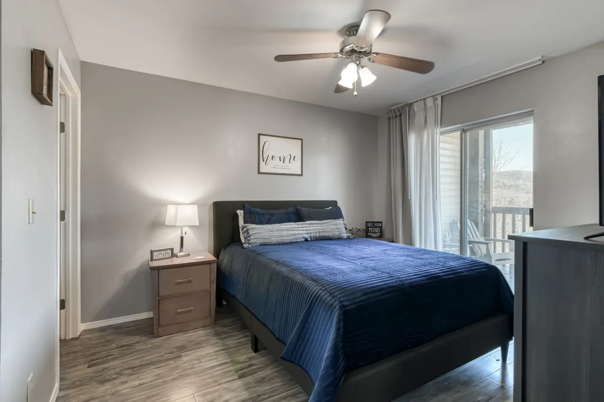 A tranquil bedroom featuring a comfortable bed made up with cozy bedding, a nightstand with a lamp, a large window allowing natural light to filter in, and a ceiling fan overhead. The space feels inviting and restful, perfect for winding down after a busy day.