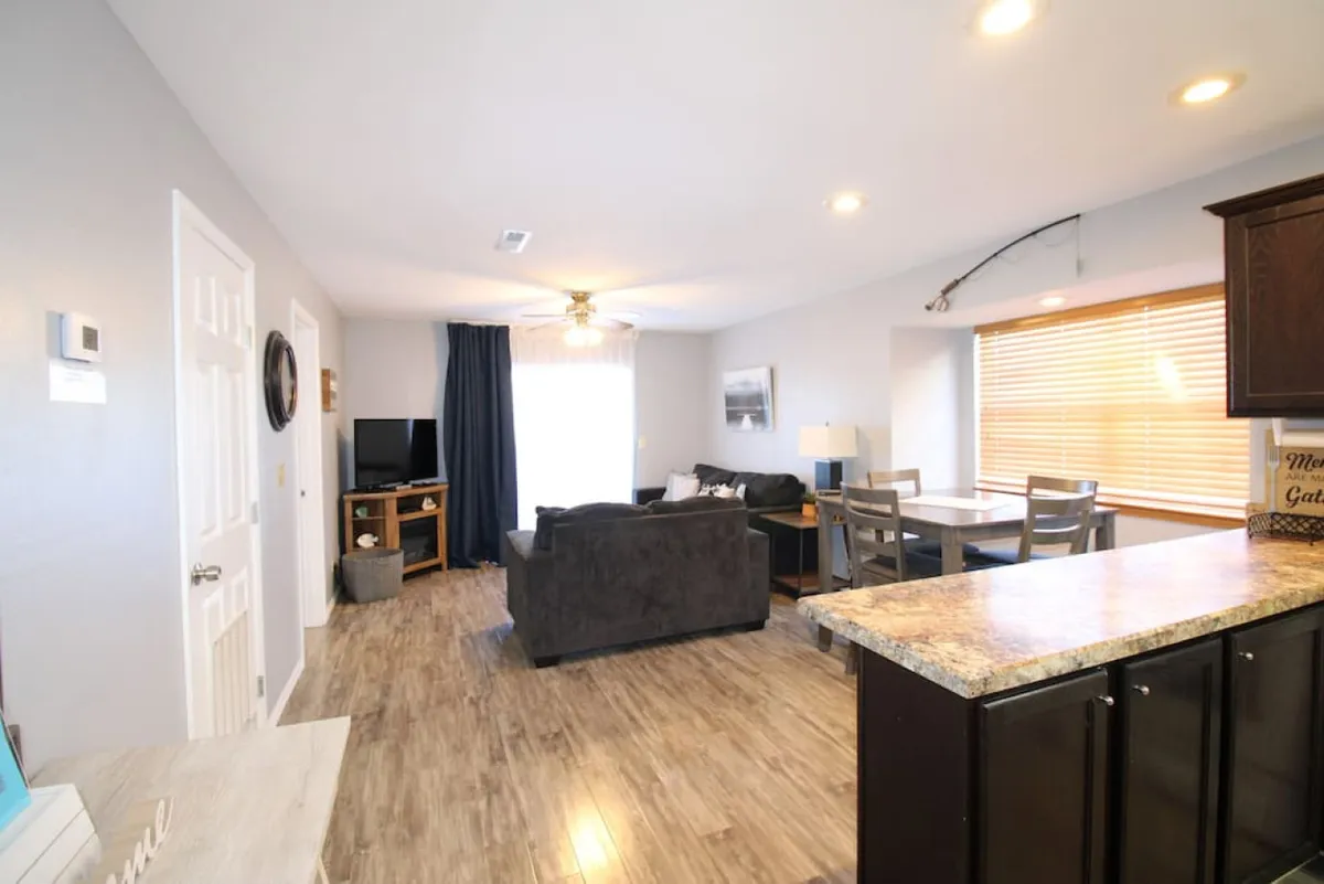 A comfortable and casual living space featuring a plush sofa, a dining table, blinds covering a large window that lets in natural light, and a television. The open-plan layout connects the living and dining area, making it ideal for relaxing, entertaining, and family meals.
