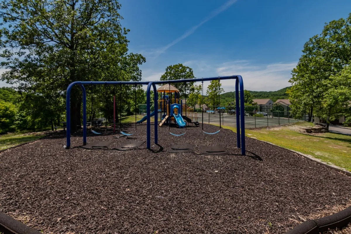 An energetic playground scene featuring modern play structures, swings, and a basketball court in the background amid lush greenery. Children are actively playing and families are gathering for picnics and games, creating a sense of community and year-round fun.