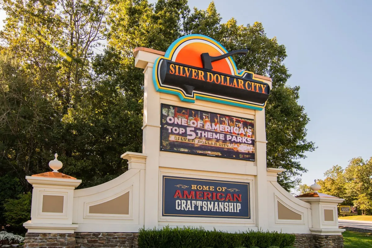A bright, cheerful exterior shot featuring the welcome sign and theme park logo at Silver Dollar City, with families gathered around the entrance. The information display adds to the sense of arrival and anticipation for a day of fun.