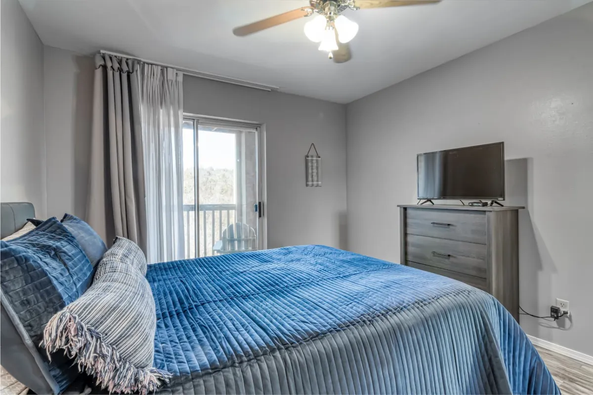 A serene bedroom with a bed dressed in a blue quilt, a large curtained window offering sunrise views, and a television set atop a dresser. The space is designed for relaxation, perfect for lazy mornings or winding down in the evening.